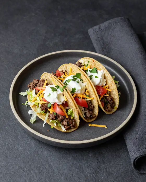 Three crunchy beef tacos in a row with lettuce, tomato, cheddar and sour cream.