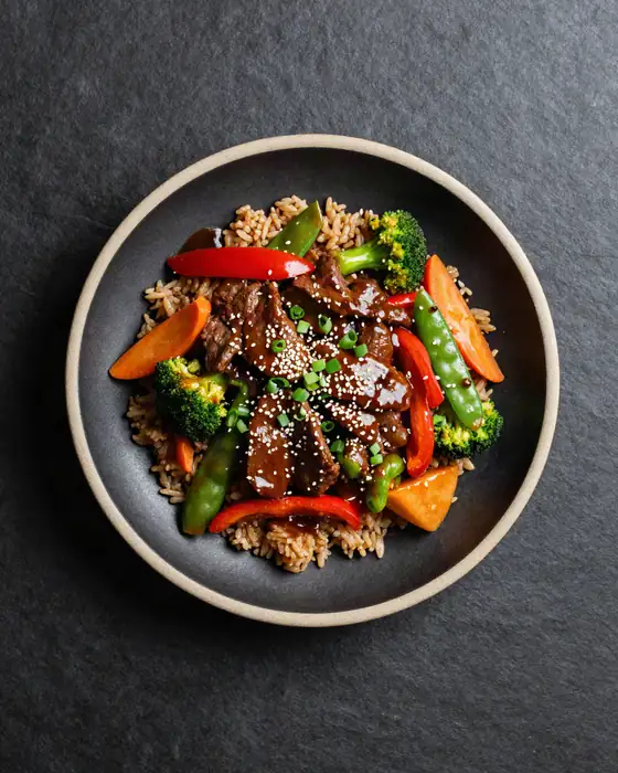 Glazed teriyaki beef with broccoli, peppers and carrots over brown rice in a dark bowl.